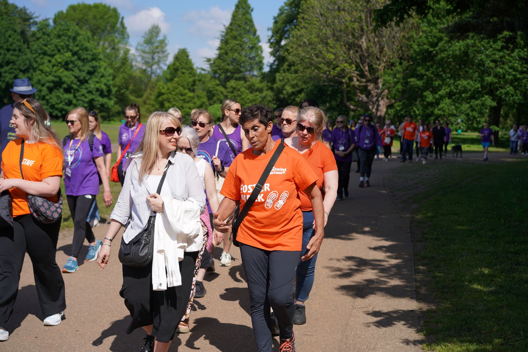 A group of people in orange and yellow t shirts walk through a park on a sunny day. Olympian Fatima Whitbread is at the front. 