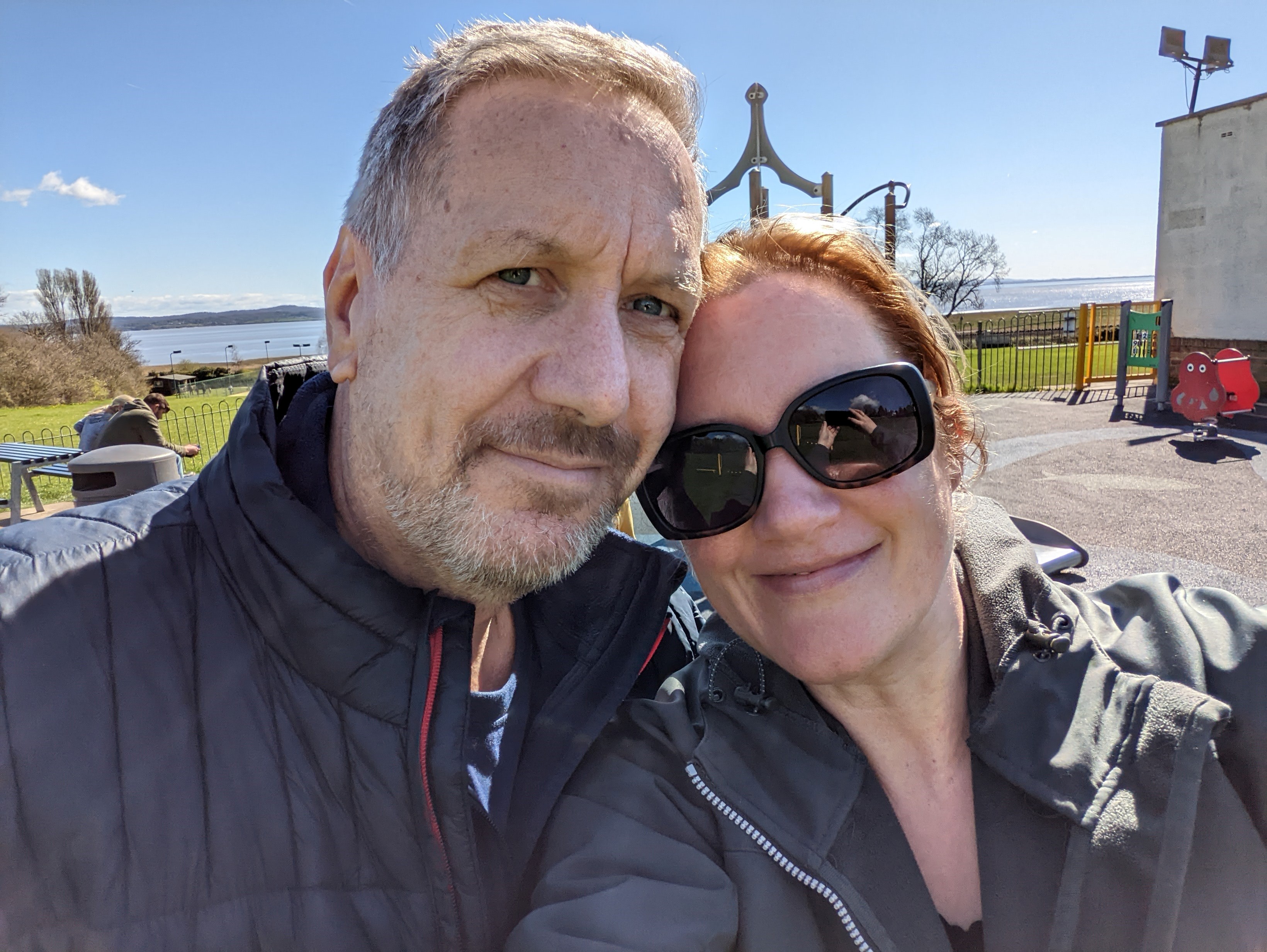 A selfie of foster carers Genevieve and Neil. They're stood in a children's play park by the sea on a gorgeous sunny day. 