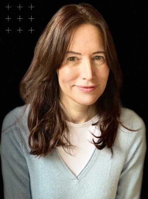 A headshot of Jess. She has got dark brown hair which falls just below her shoulders. She's wearing a light blue jumper with a white t-shirt underneath. She is in front of a black background. 