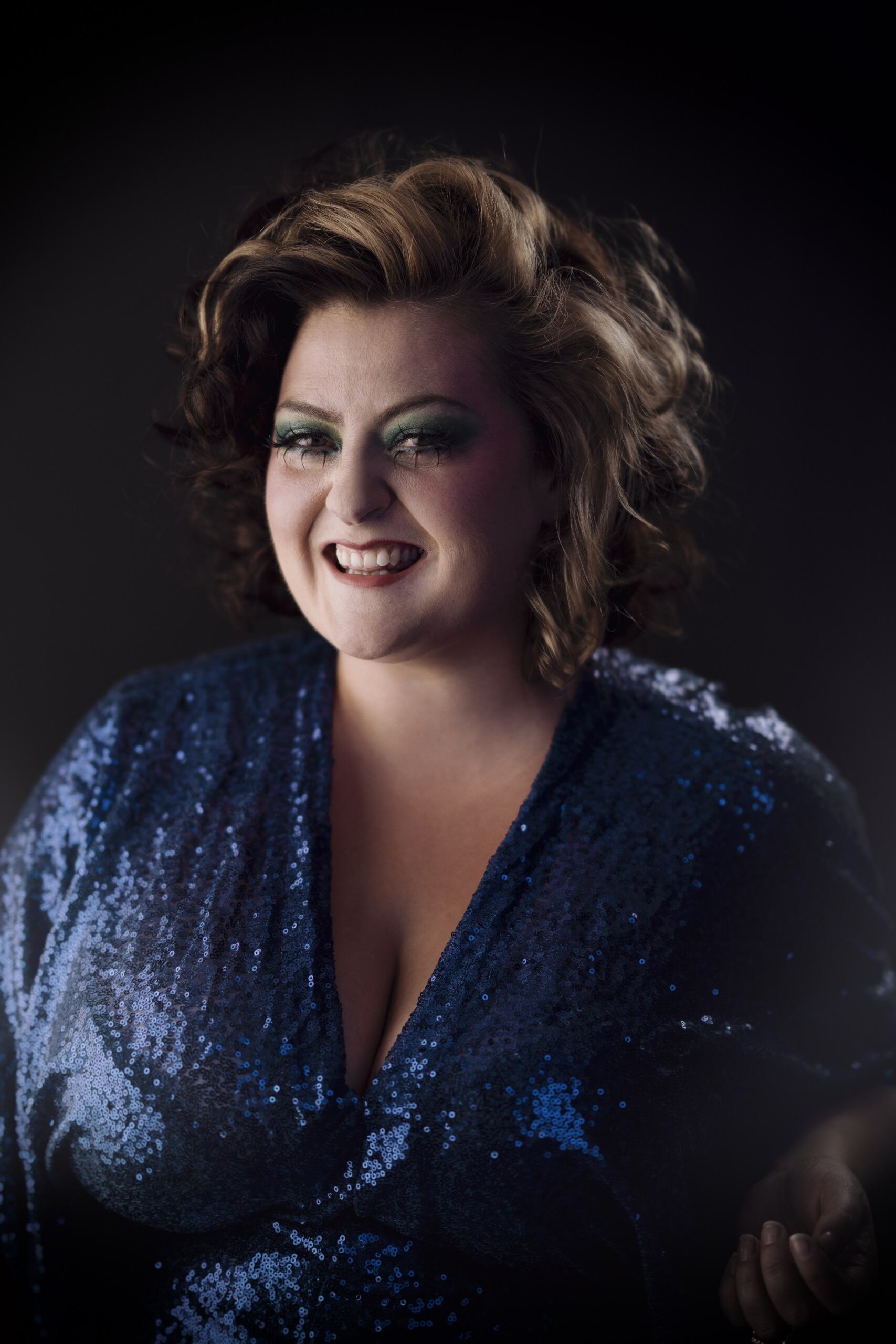 A photo of Kiri smiling at the camera. She's in front of a black background and is wearing a blue sequin dress. She has short, dark curly hair with blonde highlights. She's wearing green eyeshadow and dark red lipstick. 