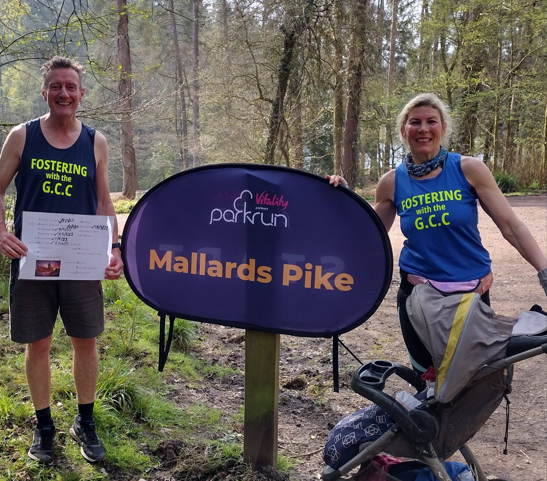 Jacqueline and John stand next to the Mallards Pike parkrun sign wearing blue running vests which state 'Fostering with the GCC' in fluorescent letters. Jacqueline is stood with the buggy, with their foster daughter out of sight so her identity is concealed. 